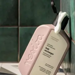 Omo jakie odżywki! Różowa butelka regenerującej odżywki do włosów "hair recharger" z pianką wydobywającą się z otwartego dozownika, na tle zielonych, łazienkowych płytek.