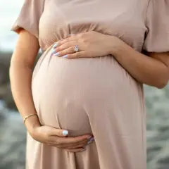 Brzuszek ciężarnej kobiety w beżowej sukience, dłonie delikatnie go obejmują. Warto pamiętać o bezpieczeństwie, np. unikaniu retinolu w ciąży.