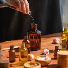 Tworzenie własnych perfum EDP. Krople płynu z pipety wpadają do bursztynowej butelki, otoczonej innymi flakonikami i suszonymi różami.