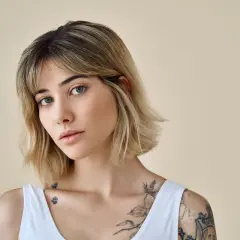 Kobieta z tatuażami i modną fryzurą long bob, z delikatnym makijażem, patrzy w obiektyw.