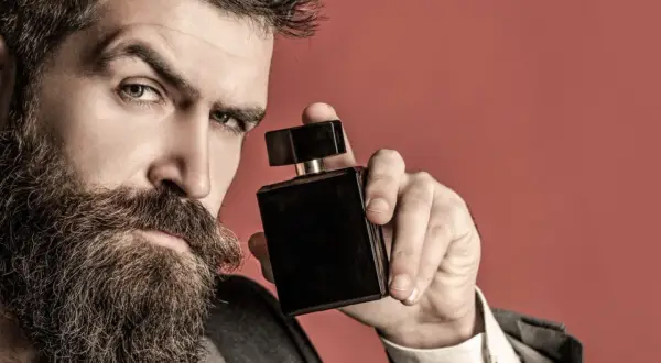 Mężczyzna z brodą trzyma czarny flakon perfum. Zastanawia się, gdzie psikac perfumami, by podkreślić swój styl.