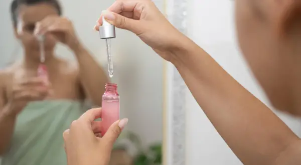 Kobieta w lustrze zastanawia się, czy retinol można stosować codziennie, trzymając w dłoni różowe serum.