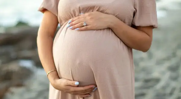Brzuszek ciężarnej kobiety w beżowej sukience, dłonie delikatnie go obejmują. Warto pamiętać o bezpieczeństwie, np. unikaniu retinolu w ciąży.