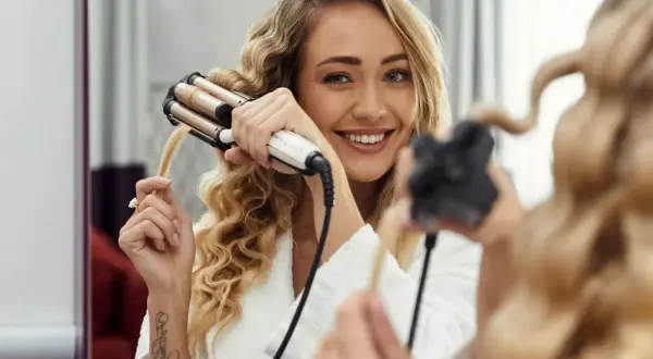 Uśmiechnięta blondynka tworzy fale na włosach do ramion za pomocą lokówki.