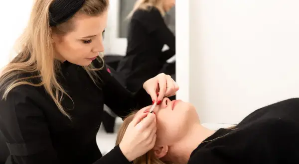 Stylistka precyzyjnie dokleja rzęsy klientce. W tle widać drugą stylistkę.