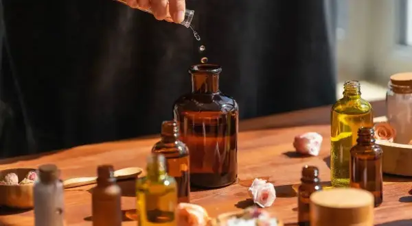 Tworzenie własnych perfum EDP. Krople płynu z pipety wpadają do bursztynowej butelki, otoczonej innymi flakonikami i suszonymi różami.