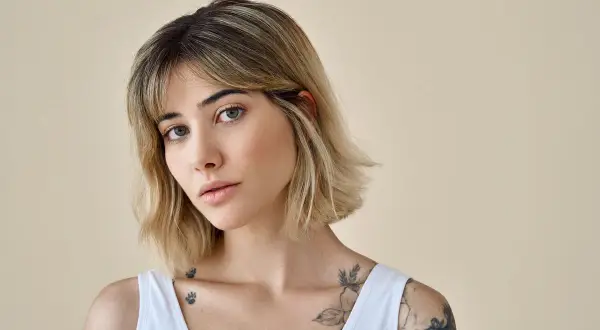 Kobieta z tatuażami i modną fryzurą long bob, z delikatnym makijażem, patrzy w obiektyw.