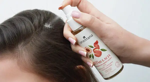 Dłoń aplikuje spray na skórę głowy, by pobudzić skręty włosów. Produkt zawiera liczi i argininę.