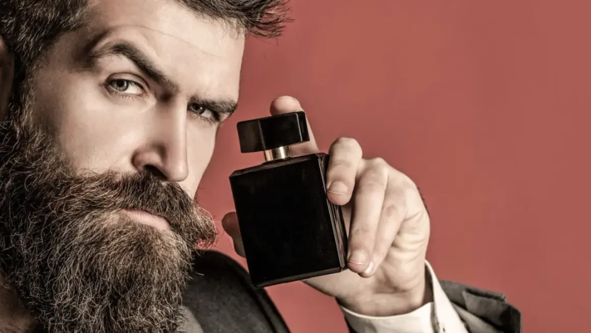 Mężczyzna z brodą trzyma czarny flakon perfum. Zastanawia się, gdzie psikac perfumami, by podkreślić swój styl.