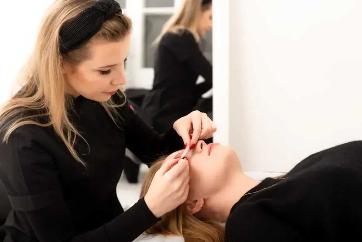 Stylistka precyzyjnie dokleja rzęsy klientce. W tle widać drugą stylistkę.