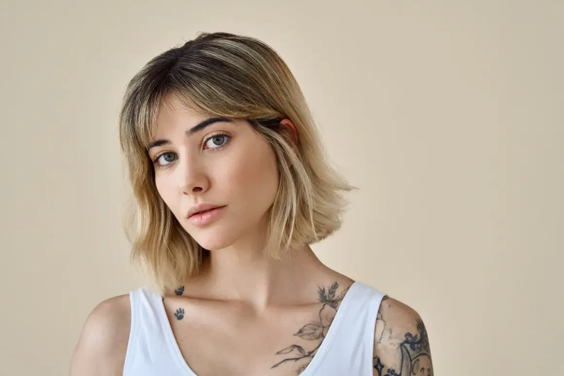 Kobieta z tatuażami i modną fryzurą long bob, z delikatnym makijażem, patrzy w obiektyw.