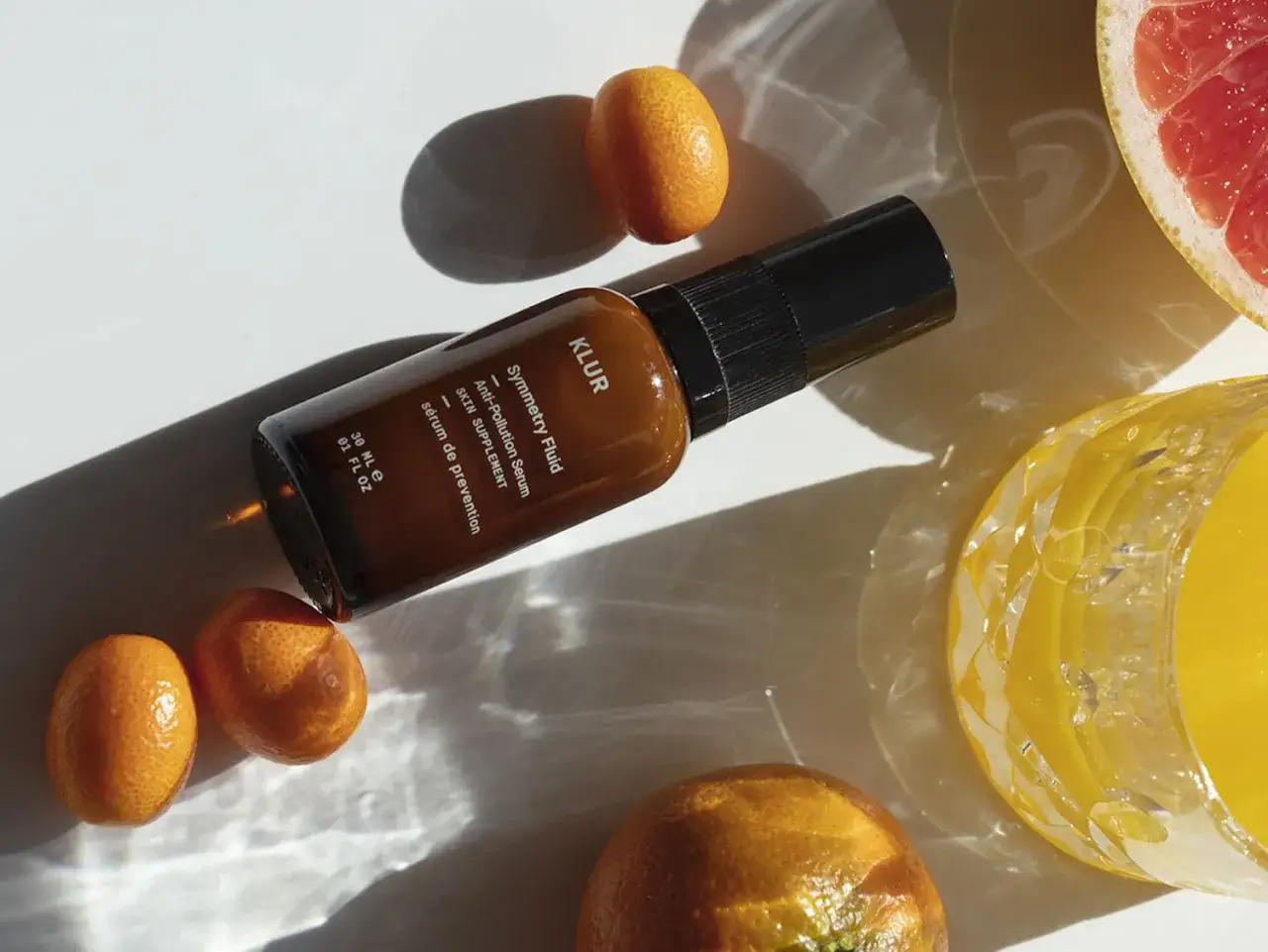 Serum KLUR Symmetry Fluid chroni sk&oacute;rę przed stresem oksydacyjnym. Wok&oacute;ł buteleczki kumkwaty, plaster grejpfruta i szklanka soku.