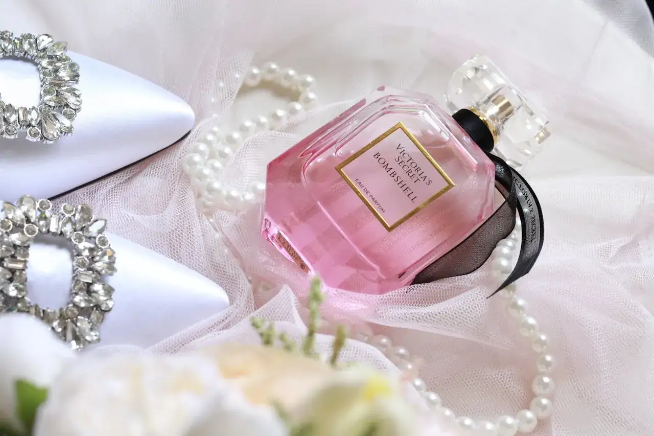 Elegancka kompozycja z r&oacute;żowym flakonem perfum Victoria's Secret Bombshell, białymi butami ozdobionymi kryształkami i perłami. Trwałość perfum oznaczenia są widoczne na etykiecie.