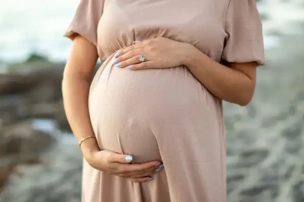 Brzuszek ciężarnej kobiety w beżowej sukience, dłonie delikatnie go obejmują. Warto pamiętać o bezpieczeństwie, np. unikaniu retinolu w ciąży.